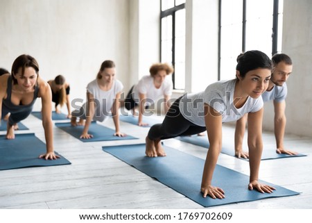 Focused young multiracial people practicing plank position on straight arms with professional indian ethnicity trainer at group class, strengthening back or abdominal core muscles together indoors.