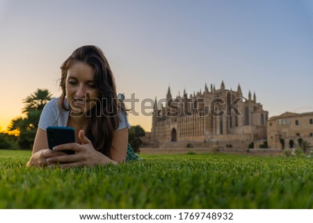 young influencer girl lying on the grass upside down using her mobile enjoying a golden sunset in front of the cathedral of palma de mallorca, brunette girl with brown hair and clear sky