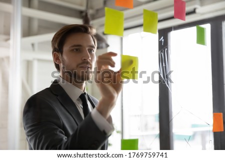 Focused young male employee write ideas on sticky notes on glass wall in office, engaged in creative thinking, motivated businessman develop company startup plan or project, brainstorm in boardroom Royalty-Free Stock Photo #1769759741