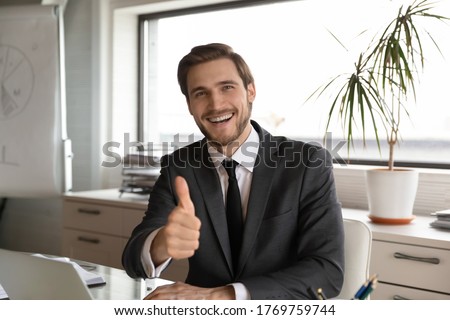 Portrait of excited young Caucasian businessman sit at desk in office show thumb up give recommendation, overjoyed male employee make like gesture advice recommend good quality company service