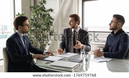 Multiracial businessmen sit at desk in office talk consider future partnership at meeting together, serious multiethnic male business partners brainstorm discuss company plan or project at briefing