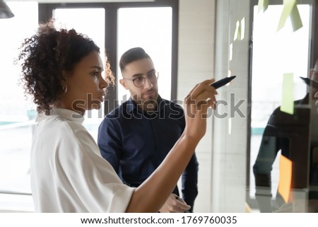 Focused multiracial employees write ideas on glass wall plan startup in office together, diverse colleagues engaged in creative thinking, develop project at meeting in boardroom, teamwork concept Royalty-Free Stock Photo #1769760035