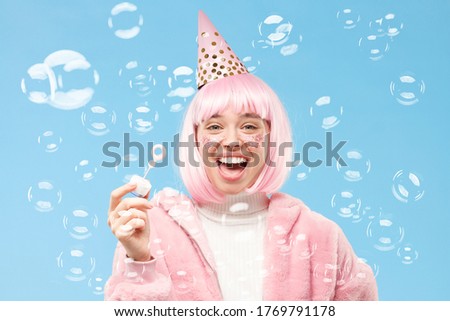 Young laughing teen girl with pink hair, coat and hat for her birthday party, blowing soap bubbles, isolated on blue background