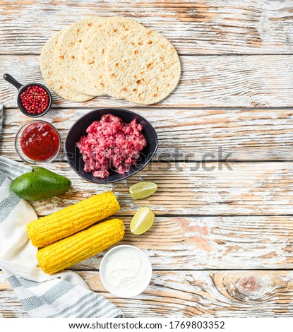 Cooking Mexican taco ingredients with minced organic beef meat in black bowl , corn, calsa over white textured wooden table, top view with space for text.