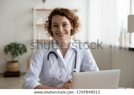Portrait of smiling young Caucasian woman nurse sit at desk in hospital or private clinic, profile picture of happy positive female doctor GP in white medical uniform at workplace, healthcare concept Royalty-Free Stock Photo #1769848013