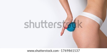 Health, people, bodycare and beauty concept. Side view of a slim fit body of a young woman doing massage on her buttocks with a massage ball with spikes on white background. Copy space