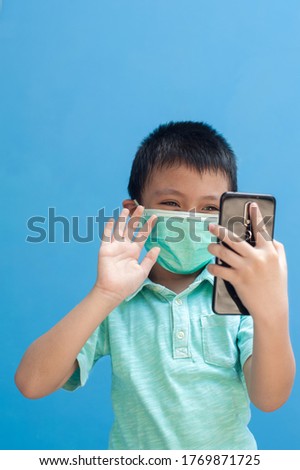 portrait of happy little boy wearing mask gesturing hi hello with his right hand to mobile phone.child communicating with other family member on video call. isolated with blue background