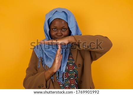Young pretty African American muslim girl tired and bored, making a timeout gesture, needs to stop because of work stress, time concept. Standing against yellow wall.
