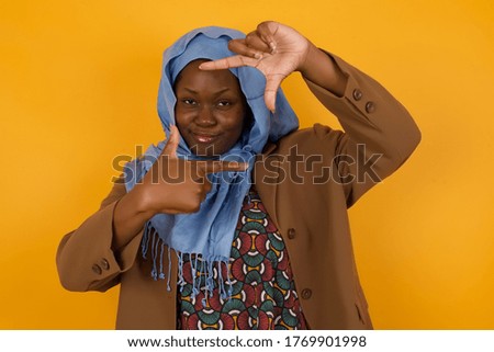 Portrait of young African American muslim positive female with cheerful expression, dressed in casual light blue sweater, has good mood, gestures finger frame actively at camera.