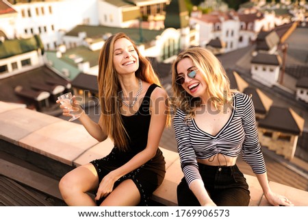 Easy-going girls with long hair expressing positive emotions in valentine's day. Excited ladies laughing and having fun. crop top and private party.
Rooftop party with the city view.Glasses and wine
