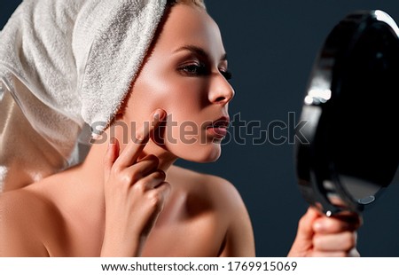 Portrait of a young attractive woman with a towel on her head looking in the mirror for acne isolated on a gray background. Skin care concept.
