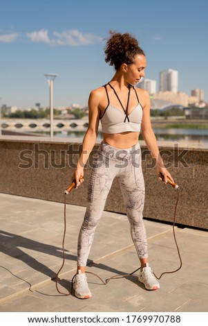 Young slim sportswoman in activewear exercising with skipping-rope while training in urban environment against riverside and blue sky