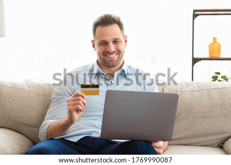 Online Shopping. Happy Man Using Laptop Holding Credit Card Purchasing Things On Internet Shop Sitting On Sofa At Home