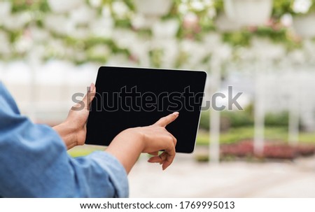 Flowers and technology. Hands of african american girl control tablet in greenhouse interior, blurred background, close up, empty screen