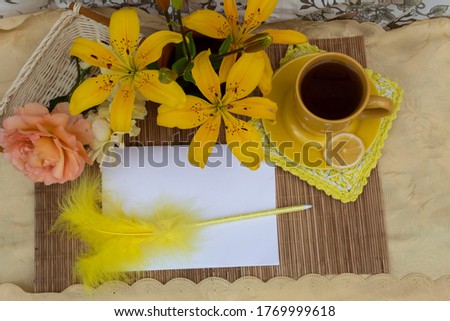 View from above. On the table is a cup of tea with lemon, a bouquet of yellow lilies and a rose. A sheet of paper lies on which lies a pen with a yellow feather.
