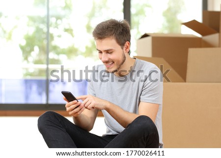 Happy man with boxes using smart phone moving home sitting on the floor surrounded by boxes