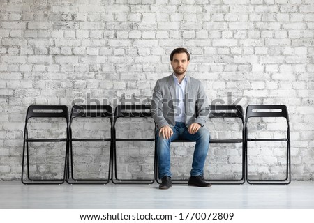 Man in jacket sit on chair alone in office hallway looking at camera feels confident and motivated hope for getting vacant place in company, prepared for job interview. HR, recruiting agency concept