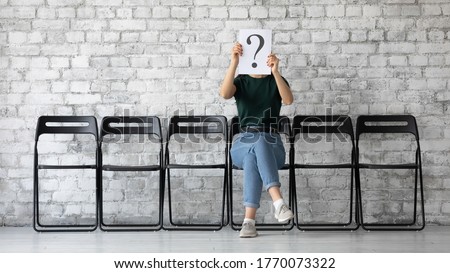 On grey brick wall background anonymous young woman sit on chair hiding her face behind paper with interrogation symbol. Doubtful female holding question mark sheet, problems and solutions HR concept Royalty-Free Stock Photo #1770073322