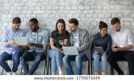 Six multi racial millennial girls and guys sit in line on chairs against grey wall using diverse devices, busy students discuss tasks, employees wait seminar in corridor having fun in internet concept Royalty-Free Stock Photo #1770073349