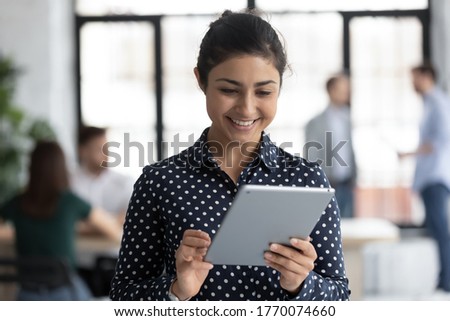 Head shot Indian millennial girl employee holding modern tablet device feels happy enjoy break at workplace having informal chat, worker browse wireless internet use pad gadget surfing web in office
