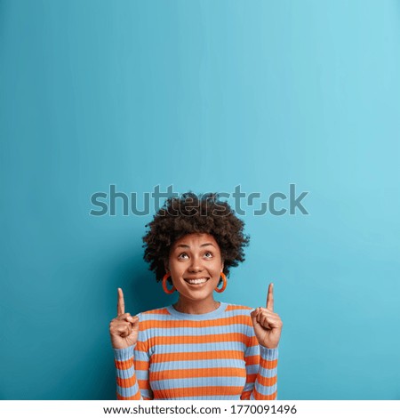 Cheerful African American woman looks up and points upwards with broad smile, recommends awesome product, suggests click banner, dressed casually, isolated on blue background shows advertisement promo Royalty-Free Stock Photo #1770091496