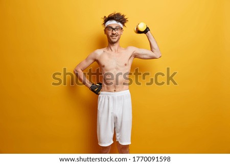 Indoor shot of positive man holds heavy sport equipment, enjoys workout in own gym, has skinny body, wants to be healthy and fit, wears transparent glasses, sport gloves, poses over yellow wall Royalty-Free Stock Photo #1770091598