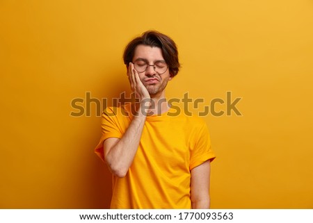 Photo of bored man looks very tired and sleepy, has dull work at day, keeps hand on cheek, closes eyes, wears round spectacles and yellow t shirt, listens to boring story, disappointed with result