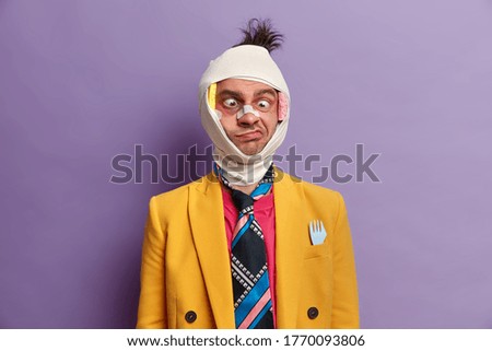 Mad crazy disabled nerdy man crosses eyes and makes funny grimace, recovers after brain concussion, being victim of bullying or assault, wears adhesive tape over broken nose, colorful formal clothing