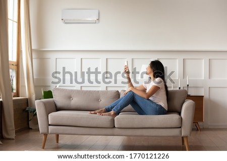 Full length smiling young asian ethnic woman sitting on cozy sofa, turning on off air conditioner with remote controller, setting indoors temperature preference, managing cooling system at home. Royalty-Free Stock Photo #1770121226