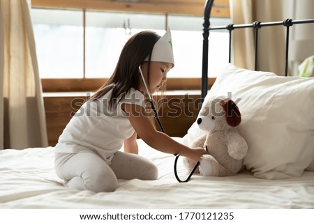 Focused adorable small vietnamese ethnicity child girl in nurse hat sitting on bed, using medical equipment, playing doctor patient checkup visit with teddy toy, imagining listening heartbeat. Royalty-Free Stock Photo #1770121235