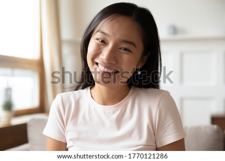 Head shot close up beautiful smiling vietnamese ethnicity young woman sitting on sofa, looking at camera. Happy biracial millennial lady blogger recording video or holding call remotely from home. Royalty-Free Stock Photo #1770121286
