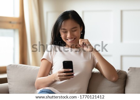 Happy sincere young asian ethnic woman looking at smart phone screen, reading sms with good news, celebrating receiving online lottery win notification, sitting on comfortable couch alone at home. Royalty-Free Stock Photo #1770121412