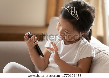Head shot interested young asian baby holding smartphone, learning using electronic device alone at home, engaged cute small vietnamese ethnicity child girl playing online video games indoors.