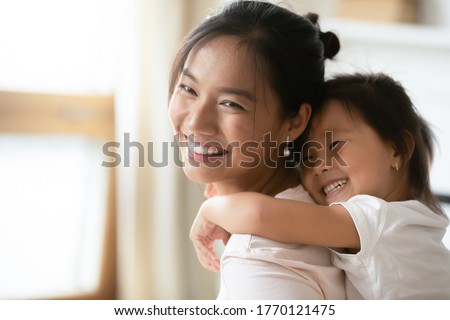 Adorable sweet little preschool kid daughter cuddling from back smiling beautiful vietnamese mother, enjoying sweet tender family moment together indoors, having fun at home, head shot close up. Royalty-Free Stock Photo #1770121475