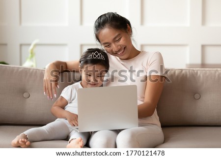 Little cute asian baby sitting on sofa with smiling affectionate vietnamese ethnicity mum nanny, looking at computer screen, watching funny childish cartoons or using early education applications.
