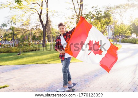 A young guy of 9 years old riding on a sunny road in the evening in the city park, dressed in a flannel plaid shirt. Holding fluttering flag of Canada. Happy Day Independence day to Canada.