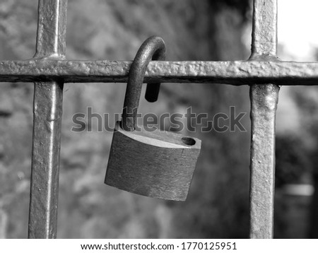 Black and white image of an old rusty open padlock hanging from an old fence. stone wall and some plants in the background.