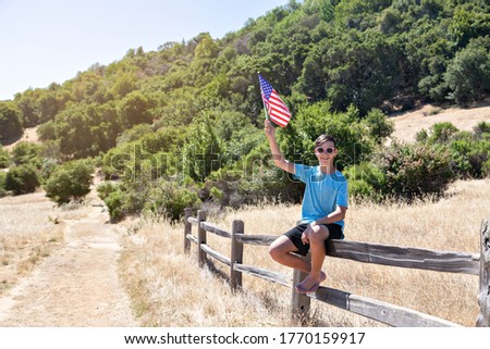 A teenager boy sits on a fence and holds the USA flag in his hand. Hiking trail in the park, summer, hot sunny day. Green background. Boy smiles. California, US