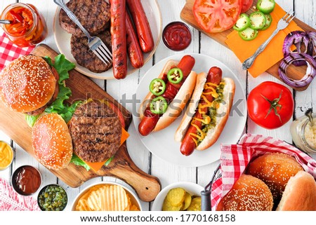 Summer BBQ food table scene with hot dog and hamburger buffet. Overhead view on a white wood background.