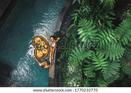 Travel happy couple in love eating floating breakfast in jungle swimming pool. Awakening in morning. Black rattan tray in heart shape, Valentines day or honeymoon surprise, view from above. Royalty-Free Stock Photo #1770210770