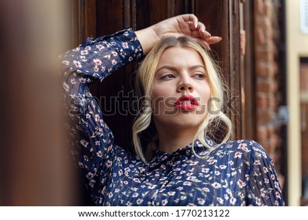 Adult caucasian blonde woman standing by the wooden door building entrance looking to the side lonely leaning wearing dress in summer day love loneliness nostalgia concept front view close up