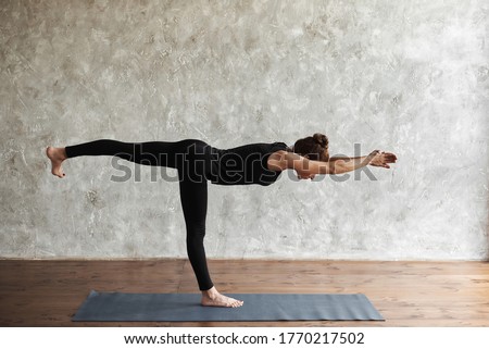 A middle-aged woman practicing yoga, doing Warrior III exercise, Virabhadrasana 3 pose, working out, wearing sportswear, black pants and shirt, indoor full length, side view. Advertising space Royalty-Free Stock Photo #1770217502