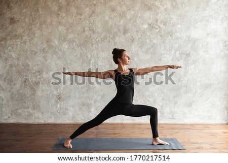 A middle-aged woman practicing yoga, doing Virabhadrasana 2 exercise, Warrior Two pose, working out, wearing black sportswear, indoor full length, grey wall in yoga studio, copy space, working out Royalty-Free Stock Photo #1770217514
