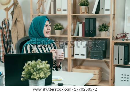 Arab woman worker in hijab holding mug and drinking coffee cup sitting in office. young islam girl employee in studio smiling enjoy city view in workplace. beautiful muslim manager relax break time