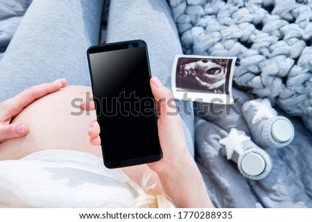 Closeup big female pregnant belly tummy. 
Ultrasound picture and little booties are on background. Young woman girl is holding smartphone with blank screen. Maternity application concept.