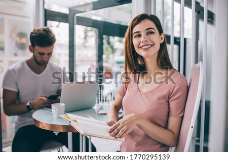 Happy modern female entrepreneur with open diary in hands smiling and looking away while sitting in cafe with male colleague