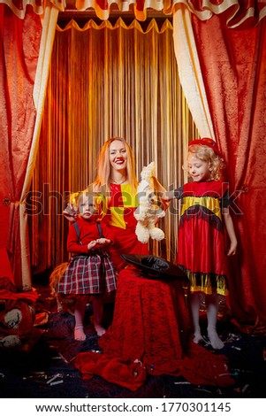 Family with dog during stylized theatrical circus photo shoot in a beautiful red location. Model mother and young daughters with small animal posing on stage with curtain
