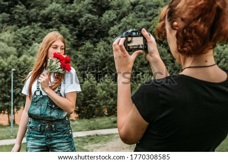 Female photographer taking photos of beautiful teen ginger girl holding bouquet of roses. Outdoors shooting, natural light