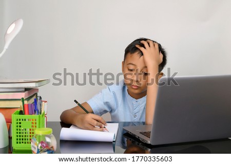 Bored, annoyed and depression of Asian schoolboy writing his homework and studying with notebook during education and learning from home for kids. Homeschooling during quarantine isolated on white.