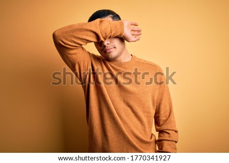Young brazilian man wearing casual sweater standing over isolated yellow background covering eyes with arm, looking serious and sad. Sightless, hiding and rejection concept